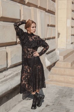 Açık havada, siyah dantelli maksi elbise giyen zarif bir kadının yaz portresi ve şehir sokaklarında poz veren sonbahar deri çizmeleri. Doğal güneş ışığı. Modelin arkasındaki şehir binaları.