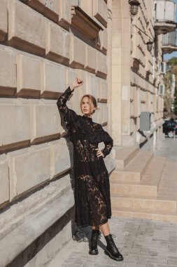 Açık havada, siyah dantelli maksi elbise giyen zarif bir kadının yaz portresi ve şehir sokaklarında poz veren sonbahar deri çizmeleri. Doğal güneş ışığı. Modelin arkasındaki şehir binaları.