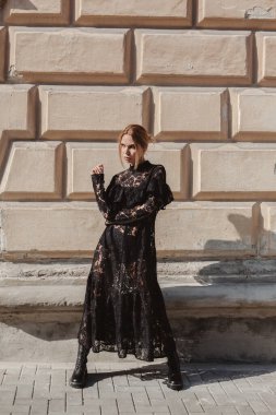 Açık havada, siyah dantelli maksi elbise giyen zarif bir kadının yaz portresi ve şehir sokaklarında poz veren sonbahar deri çizmeleri. Doğal güneş ışığı. Modelin arkasındaki şehir binaları.
