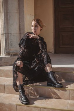 Açık havada, siyah dantelli maksi elbise giyen zarif bir kadının yaz portresi ve şehir sokaklarında poz veren sonbahar deri çizmeleri. Doğal güneş ışığı. Modelin arkasındaki şehir binaları.