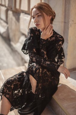 Açık havada, siyah dantelli maksi elbise giyen zarif bir kadının yaz portresi ve şehir sokaklarında poz veren sonbahar deri çizmeleri. Doğal güneş ışığı. Modelin arkasındaki şehir binaları.