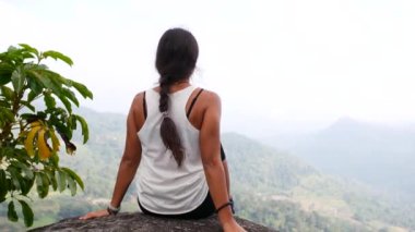Bir kız dağın tepesinde bir kayanın üzerinde oturuyor, arkadan manzaralı..