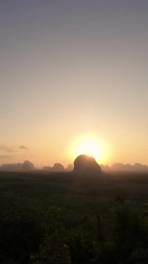 Güneş çimenli bir tarlanın üzerinde batarken dağlar çarpıcı bir zemin oluşturur. Gökyüzü renkli bulutlarla dolu, güzel doğal bir manzara yaratıyor.