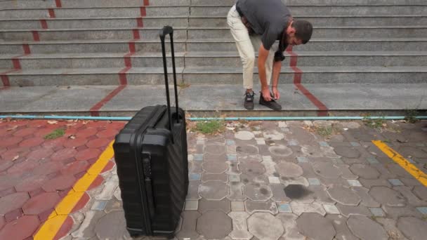 男人把他的鞋带绑在楼梯附近 前面有一个手提箱 这表明他已经准备好迎接即将到来的旅程 完美的旅行蒙太奇 包装技巧和旅行准备指南 — 图库视频影像