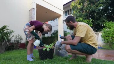 Gülümseyen bir adam ve genç bir kız fayanslı bir verandada çıplak ayakla durur, bir su bidonunu bahçe hortumuyla doldururlar. Çevrelerinde yemyeşil bitkiler ve ağaçlarla çevrili, şefkatli ve neşeli bir anı paylaşıyorlar