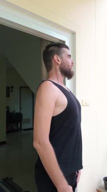 Siyah kolsuz tişörtlü bir adam kapı eşiğinde durur ve iki eliyle karnındaki yağları tutarken aşağı bakar. Yüz ifadesi memnuniyetsizlik ve öz eleştiri gösteriyor, kişisel mücadeleleri vurguluyor.