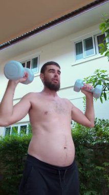 Göbeği görünen çıplak bir adam, abartılı bir kararlılıkla suratını buruşturarak başındaki hafif dambılları kaldırır. Dışarıdaki bir banliyö evinin arka planında çekilmiş.
