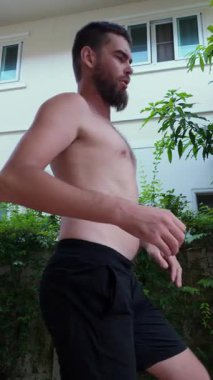 Göbeği görünen gömleksiz bir adam evin dışında duruyor, derin derin nefes alıyor ve hayal kırıklığına uğramış bir ifadeyle karnına bakıyor. Ellerini kalçalarına koyuyor, deri açıkça görülüyor.