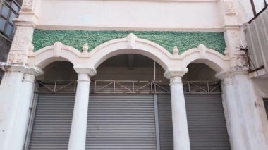 Bina cephesinde klasik sütunlar ve girişi demir kepenklerle kaplı bir kemer yer alıyor. Mimari detaylar, kentsel keşifler ve kapalı veya güvenli alanlar için uygun.