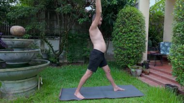 Yumuşak göbekli ve eğitimsiz bir adam arka bahçesindeki yoga minderinin üzerinde duruyor ve tereddütlü bir şekilde poz vermeye başlıyor. Yeşillik ve gündelik ortam, saf ve dürüst bir anı vurguluyor.