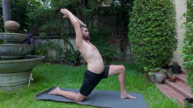 Göbeği görünen çıplak bir adam arka bahçesindeki yoga minderinin üzerinde uzanır, odaklanmış ve kararlı bir şekilde diz çökmüş vaziyette durmaya çalışır. Etrafı saksı bitkileri ve yeşilliklerle çevrili.