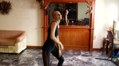 Siyah tulumlu, kel, orta yaşlı bir kadın evde dans ediyor.