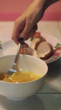 Beyaz bir seramik kasede çatal kullanarak çiğ yumurtayı çırpan bir elin yakın plan resmi. Arka planda çiçek tabağında kahverengi yumurta kabukları kırılıyor. Sahne sıcak mutfak ışığıyla aydınlatıldı.