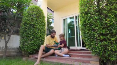 Erkek ve genç kız evlerinin dışındaki tuğla basamaklarda yan yana oturuyor, gülümsüyor ve birbirlerini yumrukluyorlar. Bahçe eldivenleri ve günlük giysiler giyerek, düzgünce budanmış