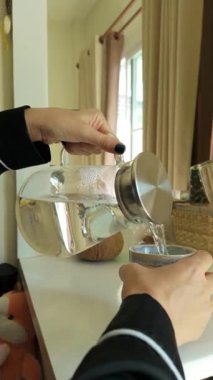 Siyah pijamalı bir kadın beyaz bir masanın yanında duruyor ve cam sürahiden seramik bir kaba tatlı su döküyor. Sabah güneşi sıcacık odayı sıcak tonlarla dolduruyor, basitliği vurguluyor ve...