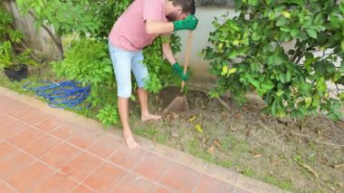 Sıradan pembe tişörtlü, kot şortlu ve yeşil eldivenli bahçedeki yaprakları tırmıklayan genç sakallı adam. Açık havaya odaklanmış, yeşil çalıların ve ağaçların yakınındaki fayanslı bir patikada yalınayak duruyor.