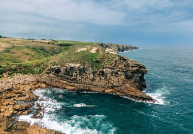 Derin mavi okyanusa bakan yeşil bitki örtüsü olan engebeli bir sahil kayalığı.