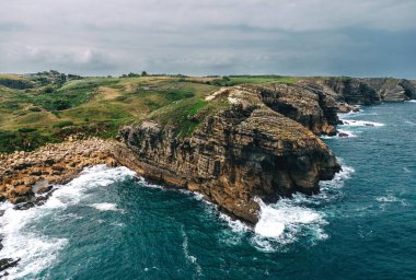 Derin mavi okyanusa bakan engebeli bir kıyı kayalığı, parçalı bulutlu bir gökyüzünün altında dalgaların kıyıya çarpması..