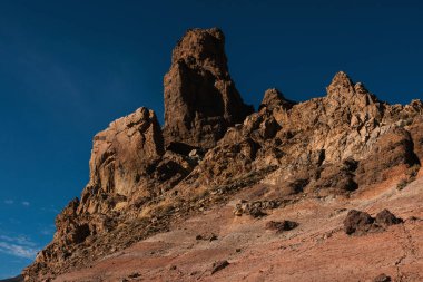 Büyük kayalık bir uçurum engebeli dokuları ve sıcak tonlarıyla derin mavi gökyüzü altında çöl benzeri bir sette.