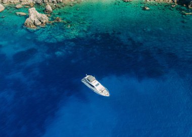 Kristal berrak mavi sularda yüzen lüks bir yatın havadan görünüşü canlı turkuaz renkli kayalık bir sahil şeridinin yanında.