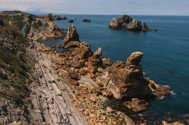 Derin mavi okyanusa uzanan katmanlı kaya oluşumları ve engebeli uçurumlarıyla dramatik bir kıyı manzarası. 