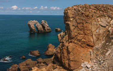 Derin mavi denizden yükselen çarpıcı bir kaya kemeriyle, dağınık bulutlu, parlak bir gökyüzünün altında devasa sahil uçurumları.