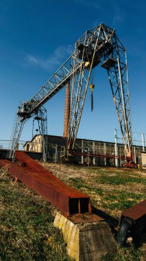 Terk edilmiş bir sanayi bölgesinde, parlak mavi gökyüzünün altında paslı kirişleri olan devasa çelik bir vinç. 