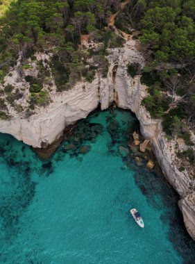 Turkuaz deniz manzaralı kayalık uçurumlar, yemyeşil orman ve kıyıya yakın yüzen bir tekne.