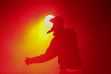 Mikrofonlu rap şarkıcısının silueti. Elinde mikrofonla havalı rapçi parlak kırmızı ışıklarla konser sahnesinde şarkı söylüyor. Hip hop sanatçısı müzikholde canlı performans sergiliyor.