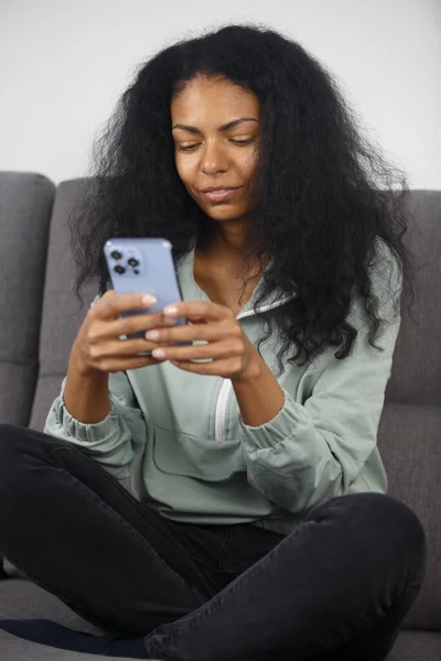 Curly black woman typing text on modern smart phone with triple camera. Adult POC female texting online with new mobile phone while sitting on couch at home