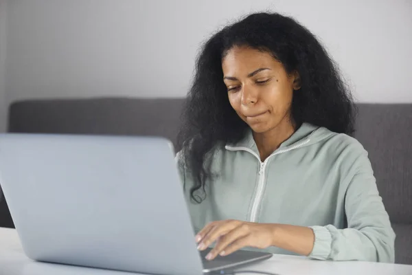 Focused black woman biting lip while typing text message on laptop keyboard. BIPOC entrepreneur female working on notebook computer at home