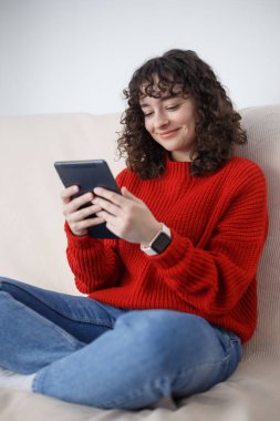 Happy Ukrainian girl reads ebook on tablet computer. Cheerful curly female person sitting on sofa at home and browsing internet on portable gadget
