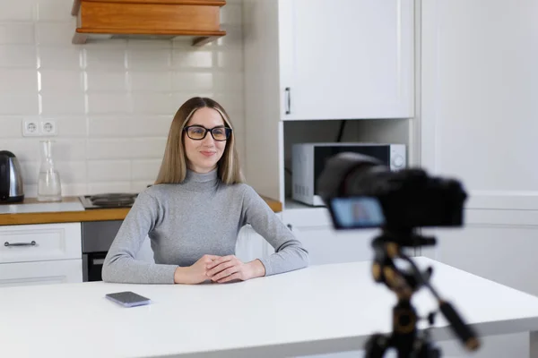 Blogger woman films video blog on professional video camera mounted on tripod. Cheerful young adult female talks on camera at home