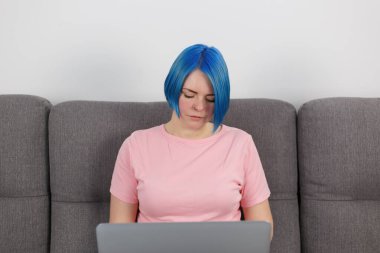 Millennial woman with dyed blue hair works on notebook computer on couch at home. Portrait of focused freelancer person working on laptop