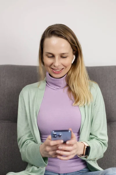 Cheerful adult woman typing sms message on smart phone. Pretty blonde female in early 40s listening to music in wireless headphones and using mobile phone app