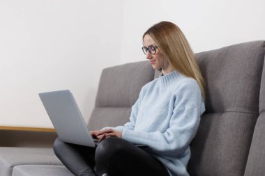 Evdeki kanepede dizüstü bilgisayarda çalışan neşeli genç bir kadın. Beyaz kadın evdeki modern dizüstü bilgisayarda yazı yazıyor.