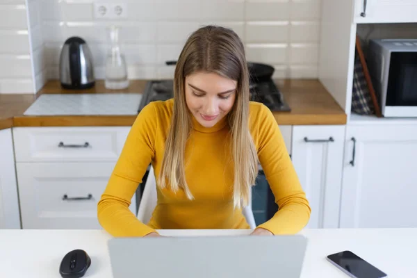Beautiful blonde woman working on modern laptop computer at home. Young adult female person typing text on notebook pc keyboard