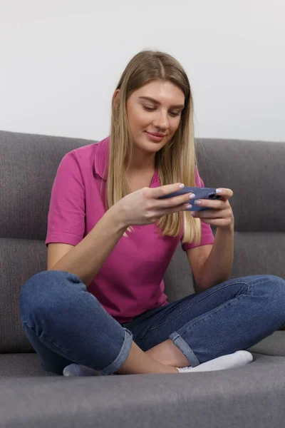 Happy blonde girl plays mobile game on smart phone. Beautiful young female person sitting on couch at home and playing online game on modern gadget