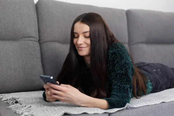 Beautiful young woman lying on couch at home and typing a message on smart phone. Cheerful brunette female texting online with modern mobile phone