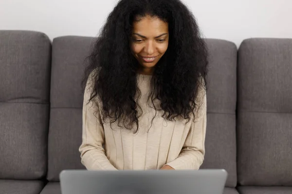 Cheerful black woman working on laptop on couch. Portrait of young adult POC female using modern notebook computer for work