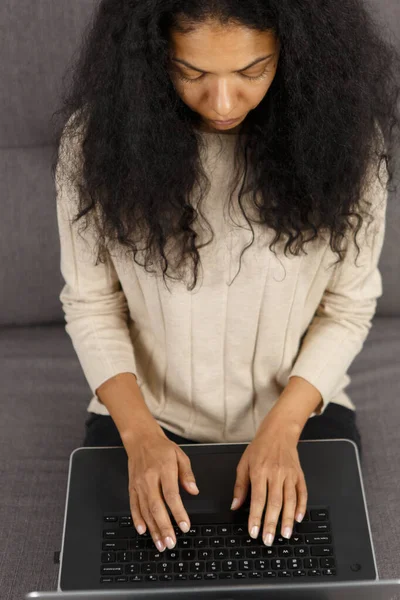 Young black woman working on laptop computer in overhead photo. Focused POC female typing text on modern notebook keyboard at home