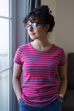 Beautiful white girl standing by the window at home and looking away. Attractive young adult female with curly hair