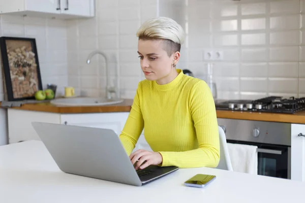 Female programmer coding on laptop. Focused white woman with short hair working on modern silver notebook pc in home kitchen