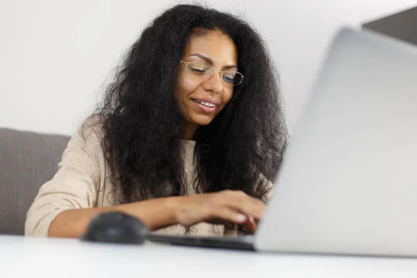 Happy black woman in glasses typing text on computer keyboard. Cheerful POC female working on modern laptop at home with a smile
