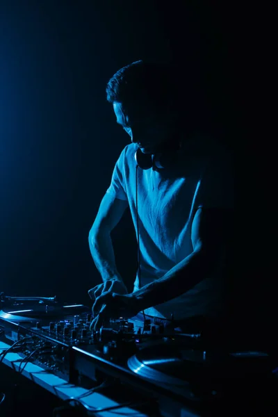 Silhouette of club DJ playing music on techno party. Disc jockey mixing musical tracks with sound mixer and vinyl records in blue light
