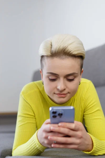 Beautiful young female lying on couch at home and typing a message on smart phone. Stylish short haired tom boy person using modern mobile phone