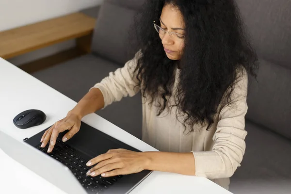 Young adult black woman working on notebook computer at home. Overhead photo of natural looking POC female typing text on laptop keyboard