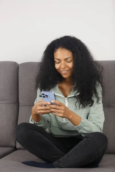 Happy black woman sitting on couch and using smart phone app. Cheerful POC female browsing mobile app on gadget with a smile