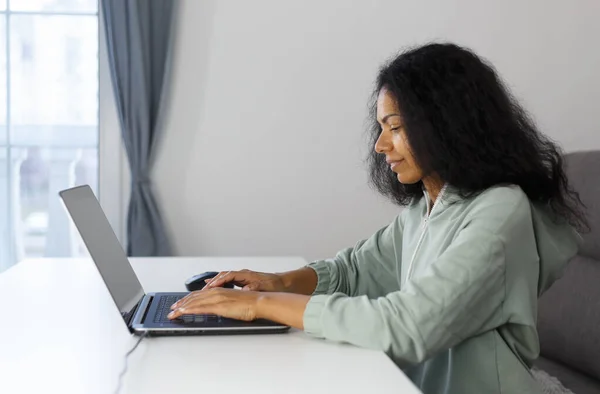 Freelance programmer coding on a laptop at home. Focused black woman typing text on modern computer with a smile