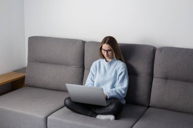 Young white woman sitting on sofa at home and working on laptop. Beautiful blonde female typing text on laptop on couch in the living room